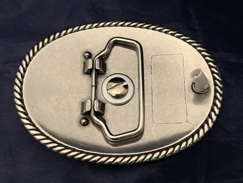 Puede incluir: Hebilla de cintur&oacute;n de plata con un borde de dise&ntilde;o de cuerda. La hebilla tiene un bucle de metal y un peque&ntilde;o pasador de metal en el lateral.