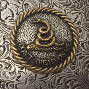 May include: A silver belt buckle with an ornate floral design. The buckle features a round, gold-colored medallion with a coiled snake and the words "Don't Tread On Me".