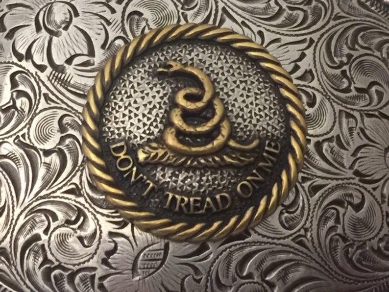 May include: A silver belt buckle with an ornate floral design. The buckle features a round, gold-colored medallion with a coiled snake and the words "Don't Tread On Me".