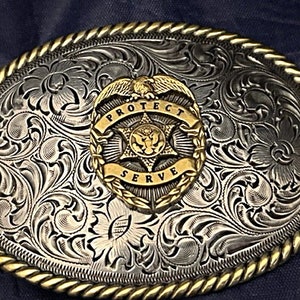 Puede incluir: Hebilla de cintur&oacute;n de plata con un dise&ntilde;o de &aacute;guila y estrella dorado. La hebilla tiene un patr&oacute;n floral grabado y las palabras "Protect Serve" alrededor del &aacute;guila.
