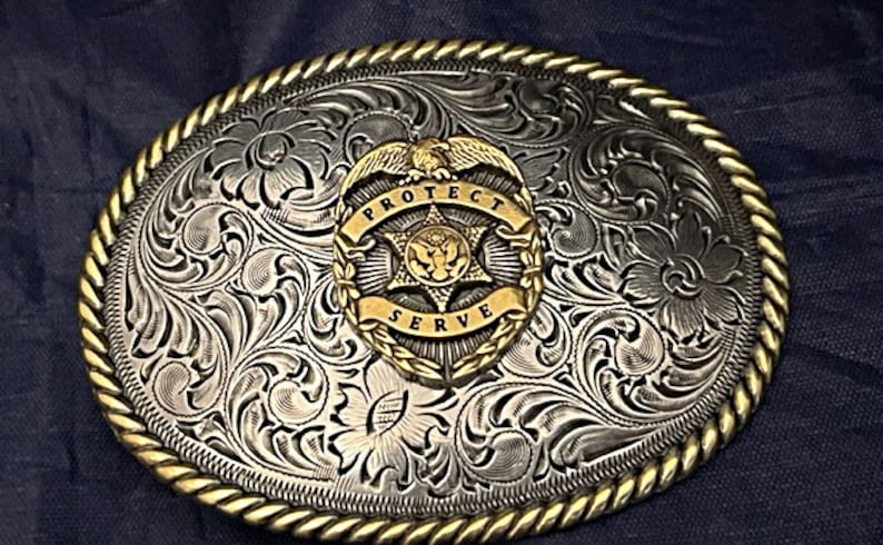 Puede incluir: Hebilla de cintur&oacute;n de plata con un dise&ntilde;o de &aacute;guila y estrella dorado. La hebilla tiene un patr&oacute;n floral grabado y las palabras "Protect Serve" alrededor del &aacute;guila.