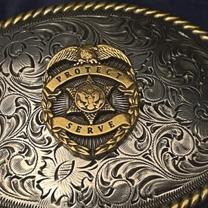 Puede incluir: Hebilla de cintur&oacute;n de plata y oro con un dise&ntilde;o floral ornamentado. En el centro, una insignia dorada con un &aacute;guila, una estrella y las palabras "Protect Serve".