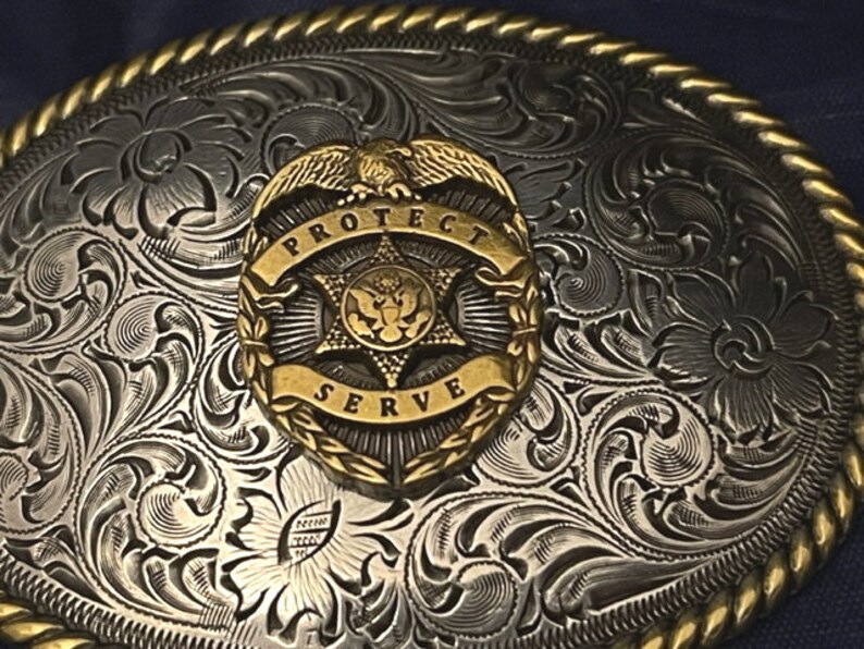 Puede incluir: Hebilla de cintur&oacute;n de plata y oro con un dise&ntilde;o floral ornamentado. En el centro, una insignia dorada con un &aacute;guila, una estrella y las palabras "Protect Serve".