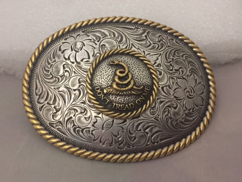 May include: A silver and gold belt buckle with an engraved floral design. The center of the buckle features a gold snake with the words "Don't Tread On Me".