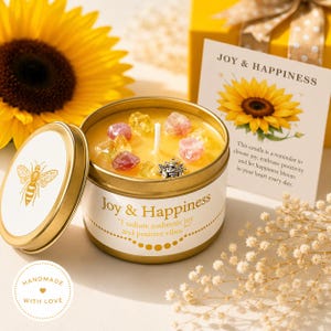 May include: A gold-toned metal candle tin labeled Joy & Happiness with a bee illustration on the lid. The yellow wax is topped with small colorful crystals and a silver bee charm. A matching card features a sunflower and the text Joy & Happiness.