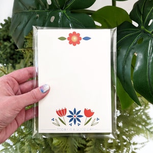 Op de afbeelding: Notitieblok met een bloemenontwerp in een doorzichtige plastic hoes. Het notitieblok heeft een crèmekleurige achtergrond met rode, blauwe en groene bloemaccenten. De tekst "TODAY IS A GOOD DAY" staat onderaan.