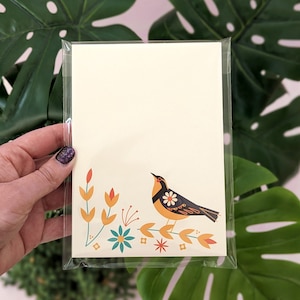 May include: A cream-colored stationery set featuring a stylized bird design with orange, black, and white accents. The bird is perched on a branch with floral elements in orange, teal, and red. The stationery is packaged in a clear plastic sleeve.