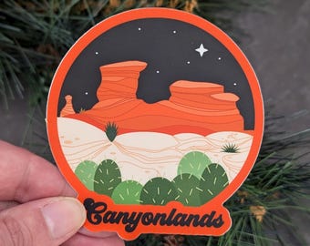 May include: A round sticker with a retro design featuring a red rock formation under a starry night sky. The sticker has the text "Canyonlands" in a retro font.