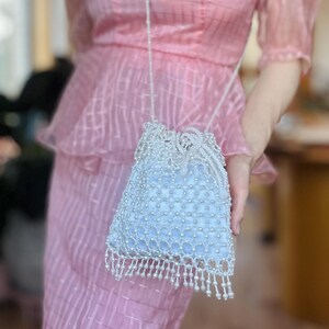 Può includere: Una borsa a sacchetto azzurra ricoperta di perline e perle bianche, con frange di perline sul bordo inferiore e una sottile tracolla di perline. La borsa è tenuta davanti a un abito rosa testurizzato con maniche a sbuffo e baschina.