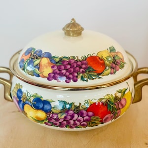 May include: Cream-coloured enamel casserole dish with lid, decorated with a colourful fruit design including grapes, apples, pears, and plums. The dish has gold-toned handles and trim, and a gold finial on the lid. A decorative kitchenware item.