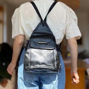 May include: A black backpack with a triangular top flap and a gold-toned metal plaque featuring the text ITALIA. The bag has a textured finish, thin shoulder straps, and a main compartment secured with a flap closure and visible zipper hardware.
