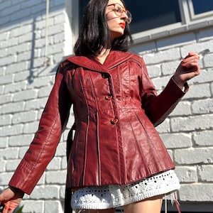 May include: A dark red leather jacket featuring a wide collar, long sleeves, and a button-front closure. The jacket has vertical paneling and a cinched waist design. It is paired with white crochet shorts that have a scalloped hemline detail.