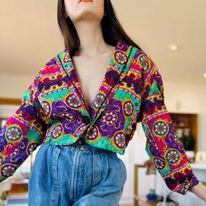 May include: A cropped long-sleeve jacket featuring a vibrant, multi-coloured baroque-inspired pattern in shades of purple, bright pink, green, and gold, with a single black button closure and black piping along the lapels and front edges.
