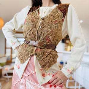 May include: A cream-colored long-sleeve cardigan with floral embroidery on the cuffs and hem, layered under a brown textured vest with intricate stitching and button closures. A brown braided leather belt with a metal buckle cinches the waist.