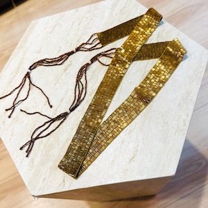 May include: A long, narrow belt crafted from metallic gold seed beads arranged in a rectangular grid pattern. The belt features long, thin brown beaded ties at each end, accented with small, light-colored spherical beads along the length of the ties.