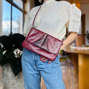 May include: A deep burgundy leather shoulder bag featuring a rectangular silhouette, a fold-over flap closure, and a central panel with a textured, snakeskin-like embossed pattern. The bag includes a thin, matching leather shoulder strap.