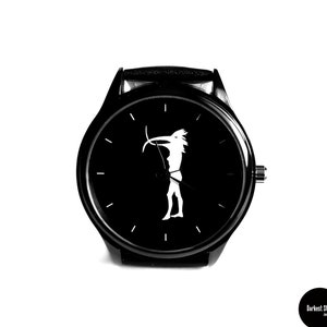 Może przedstawiać: Czarny zegarek na rękę z czarnym skórzanym paskiem i białą sylwetką osoby z łukiem i strzałą na tarczy.