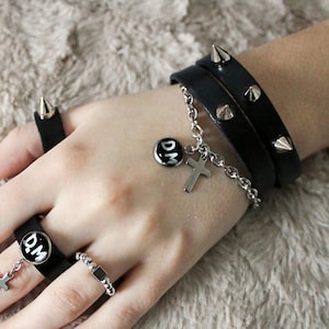 Może przedstawiać: Ręka z kilkoma srebrnymi pierścionkami z czarnymi akcentami i czarną skórzaną bransoletką z srebrnymi ćwiekami i srebrnym wisiorkiem z literami "D.M." i krzyżem. Ręka pomalowana jest na czerwono.