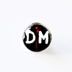 May include: A black and white circular pin with the letters "DM" in white and a red vertical line in the centre. The pin is on a white background.