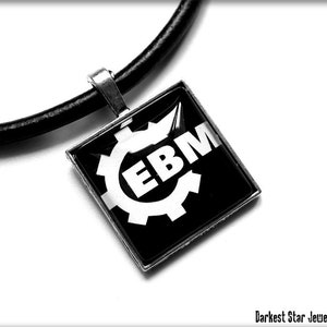 May include: Black and white pendant necklace with a gear and the letters EBM on a black background. The pendant is attached to a black leather cord.