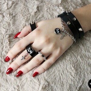 May include: A hand wearing a black leather bracelet with silver studs and a silver chain with a cross charm and a round charm that says "D.M.". The hand is also wearing several silver rings, including one with a black band and a round charm that says "D.M.". The hand has red nail polish.