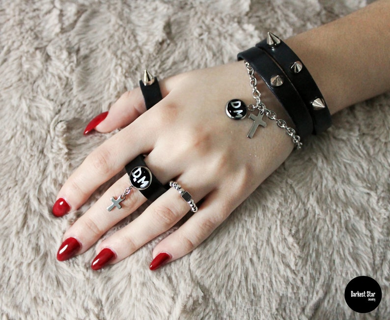 May include: A hand wearing a black leather bracelet with silver studs and a silver chain with a cross charm and a round charm that says "D.M.". The hand is also wearing several silver rings, including one with a black band and a round charm that says "D.M.". The hand has red nail polish.