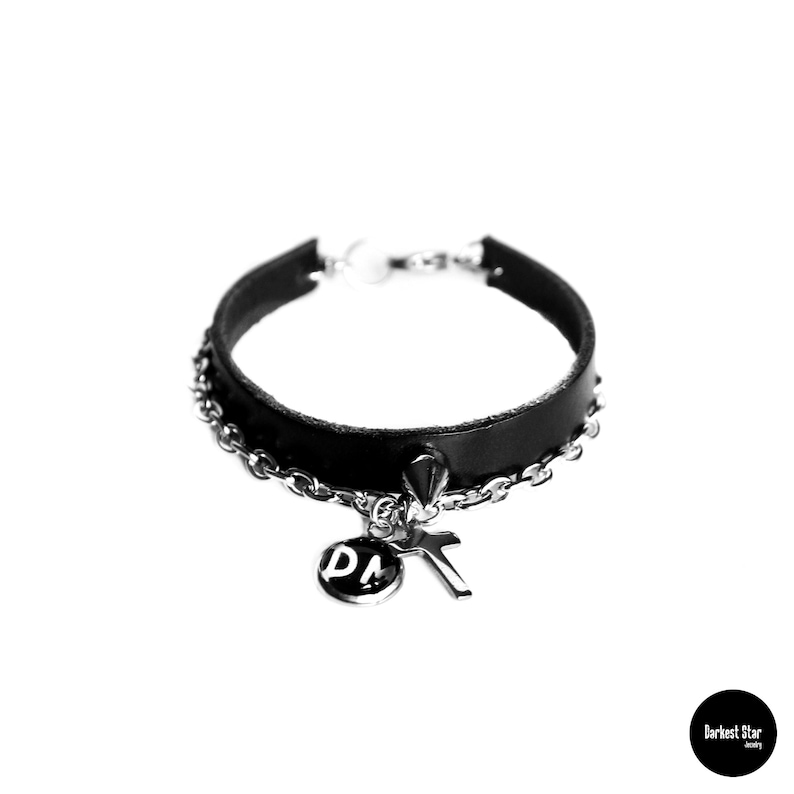 May include: Black leather bracelet with a silver chain and two charms: a cross and a round charm with the letters "DM".
