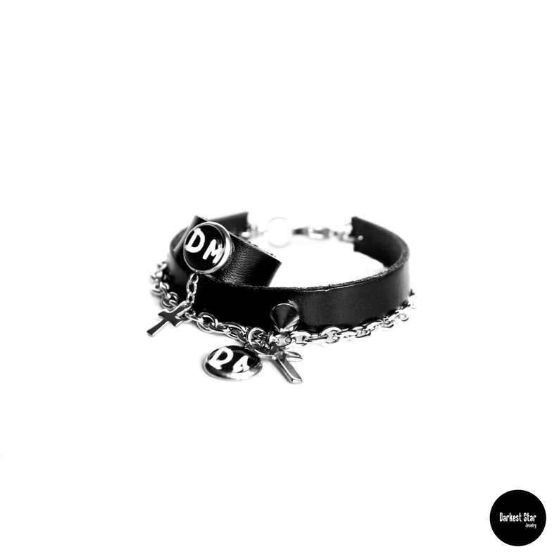 May include: Black leather bracelet with a silver chain and two silver charms. One charm is a circle with the letters "DM" in white, and the other is a cross.