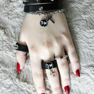 May include: A person's hand wearing a black leather bracelet with silver studs, a silver chain bracelet with a cross charm, and a silver ring with a black 'DM' charm. The person is also wearing a silver ring with a black 'DM' charm and a silver ring with a small silver rectangle charm. The person's fingernails are painted red.