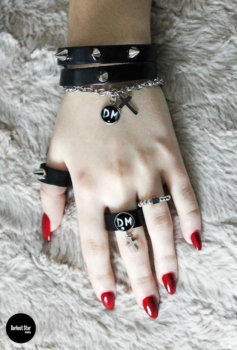 May include: A person's hand wearing a black leather bracelet with silver studs, a silver chain bracelet with a cross charm, and a silver ring with a black 'DM' charm. The person is also wearing a silver ring with a black 'DM' charm and a silver ring with a small silver rectangle charm. The person's fingernails are painted red.