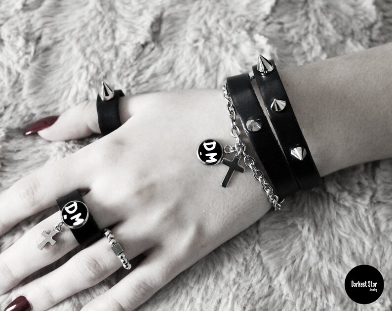 May include: A black leather bracelet with silver studs and a silver chain with a cross charm and a round charm with the letters "DM" in white. The bracelet is worn on a person's wrist.