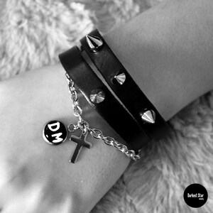 May include: A black leather bracelet with silver studs and a silver charm with the letters "DM" on it. The bracelet is worn on a person's wrist.