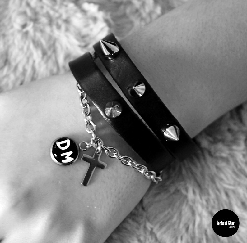 May include: A black leather bracelet with silver studs and a silver charm with the letters "DM" on it. The bracelet is worn on a person's wrist.