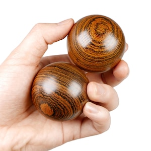 Puede incluir: Dos bolas de madera marrón con un patrón rayado se sostienen en la mano de una persona. Las bolas se utilizan para el ejercicio y la relajación.