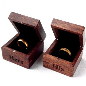 Könnte beinhalten: Zwei hölzerne Ringboxen mit geöffneten Deckeln, die goldene Ringe im Inneren zeigen. Die Boxen sind mit den Worten "Hers" und "His" graviert.