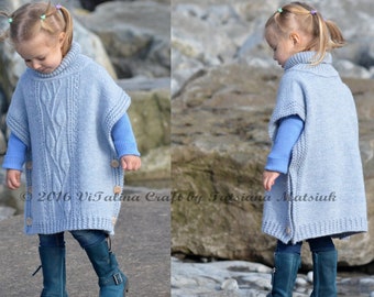 May include: A light blue knitted poncho with a cable knit pattern and wooden buttons. The poncho is worn by a child standing on a rocky beach.