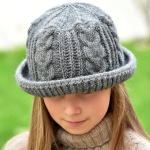 Puede incluir: Un sombrero cloche gris tejido con un diseño de punto trenzado y un ala pequeña. El sombrero se lleva con un jersey de cuello alto beige. El fondo es verde borroso.