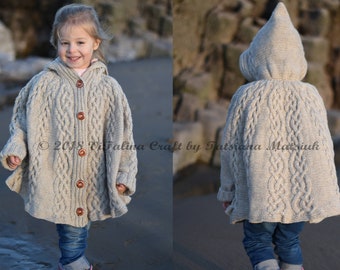 May include: A young child wearing a beige knitted hooded cape with a cable knit pattern and wooden buttons. The cape is worn over blue jeans and pink rain boots. The child is standing on a sandy beach.