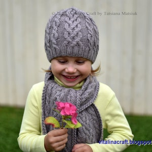May include: A young person wearing a gray knitted hat and scarf with a cable knit pattern. They are holding a pink flower.