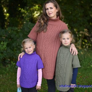 May include: A woman and two girls wearing matching cable knit sweaters in different colors. The woman is wearing a rust colored sweater, the older girl is wearing a green sweater, and the younger girl is wearing a purple sweater. All three sweaters have a high neck and short sleeves.