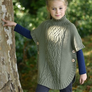 May include: A young person wearing a green cable knit poncho with wooden buttons. The poncho has a high neck and is worn over a blue long-sleeved shirt and dark blue jeans.