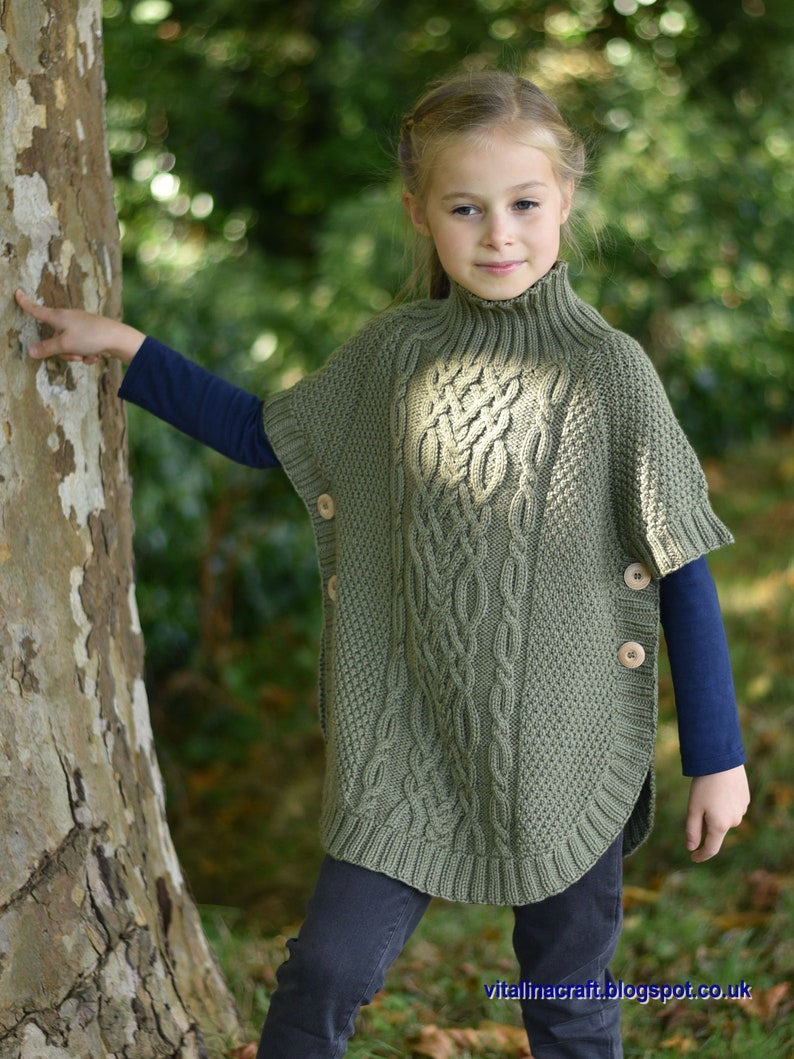 May include: A young person wearing a green cable knit poncho with wooden buttons. The poncho has a high neck and is worn over a blue long-sleeved shirt and dark blue jeans.