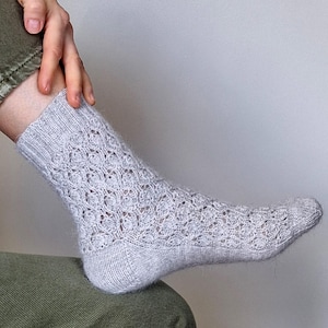 May include: A light gray knit sock featuring a delicate, repeating openwork lace pattern across the foot and ankle, with a ribbed cuff at the top and a smooth, solid knit texture on the heel and toe sections, crafted from a soft, fuzzy yarn.
