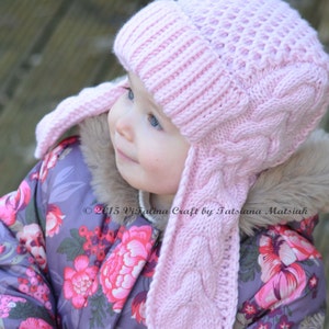 May include: A light pink knitted hat with ear flaps. The hat features a cable knit design on the ear flaps and sides, with a textured pattern on the crown. The hat is worn by a child wearing a floral print jacket.