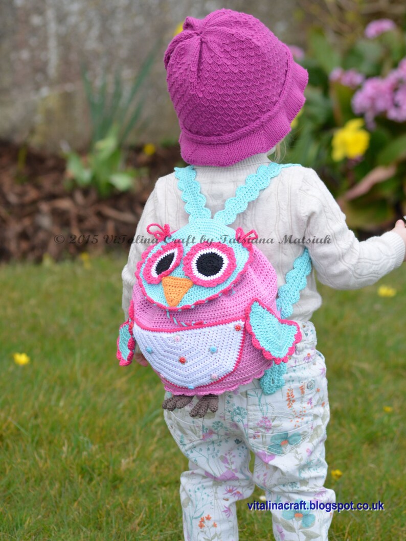 May include: A pink and blue crocheted owl backpack with a white and pink patterned belly. The backpack is being worn by a child wearing a pink hat and white pants with a floral pattern.
