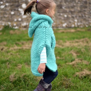 May include: A young girl wearing a teal knitted hooded poncho with a white striped shirt underneath. She is standing in a grassy field with a stone wall in the background.