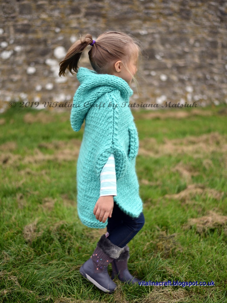 May include: A young girl wearing a teal knitted hooded poncho with a white striped shirt underneath. She is standing in a grassy field with a stone wall in the background.