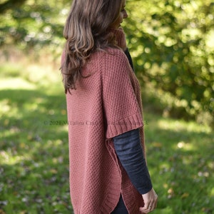 K&ouml;nnte beinhalten: Eine Frau tr&auml;gt einen rostigen Strickponcho mit hohem Kragen und gerippter Struktur. Der Poncho hat eine leicht ausgestellte Form und wird &uuml;ber einem schwarzen Langarmshirt und einer schwarzen Hose getragen.