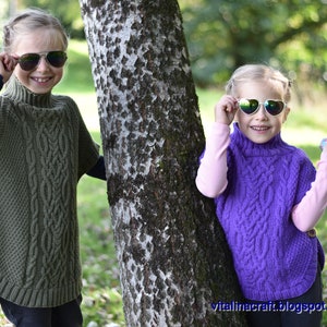 May include: Two children wearing knitted ponchos with cable knit patterns. The girl on the left is wearing a green poncho and the girl on the right is wearing a purple poncho. Both girls are wearing sunglasses.