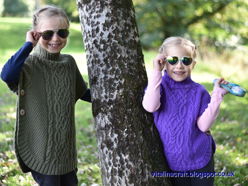 May include: Two children wearing knitted ponchos with cable knit patterns. The girl on the left is wearing a green poncho and the girl on the right is wearing a purple poncho. Both girls are wearing sunglasses.